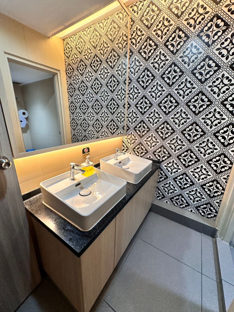 Two rectangular white sinks set into a dark granite countertop under a wide wall mirror in a bathroom with black-and-white patterned tile walls and warm under-cabinet lighting.