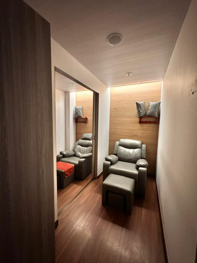 Narrow spa or salon treatment room with two gray leather armchairs and matching ottomans, wood-paneled walls, and soft lighting.
