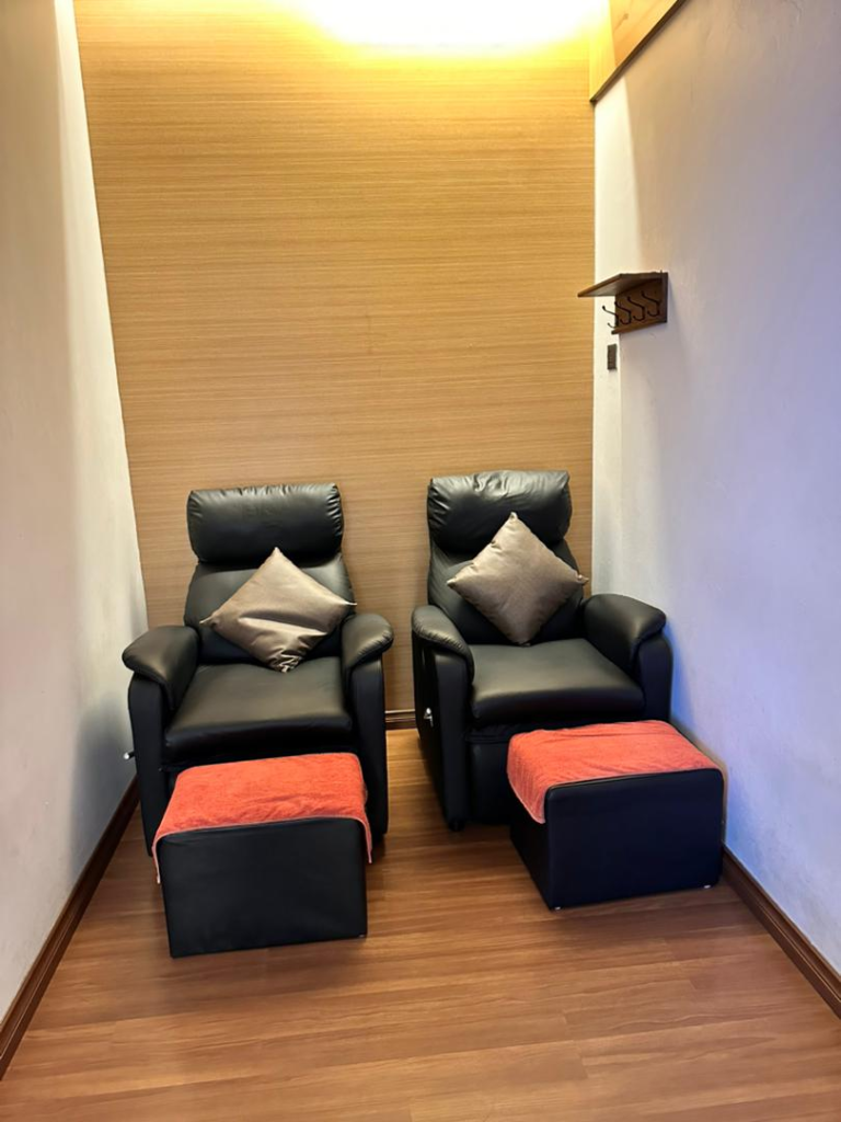 Pair of black leather armchairs with matching ottomans and peach pillows in a narrow wooden-walled relaxation room, towels folded on ottomans.